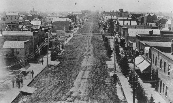 Elevated view looking south along unpaved Tower Avenue of storefronts, American flags are flying from a number of the commercial buildings.