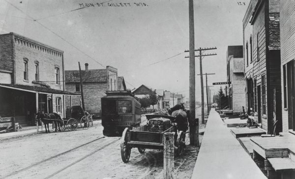 View from sidewalk on the right down a street, with horse-drawn wagons parked on the left and right curbs, and a streetcar in the center. A sign on the right over the sidewalk reads: "Bakery." A note on the reverse of the photograph reads: "Sorenson store at extreme left."