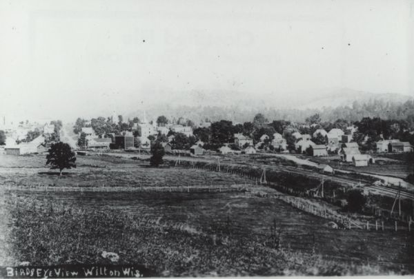 Slightly elevated view of a town, looking down from a hill across a field, with railroad tracks curving from the right foreground to the left towards the town in the middle distance. Forests and hills are in the far distance behind the town.
