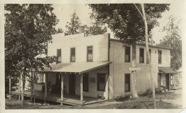 View of a building with a covered porch, and a sign on a tree that reads: "Boats to Let." A group of people are sitting on the porch. There is a body of water in the background.
