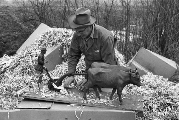 A man in hat and glasses is unpacking boxes. In the foreground on top of a box is a sculpture of a plowing scene. Original caption reads: "17th-century plow is unwrapped by Curator Edward D. Carpenter. Stonefield was selected from 32 U.S. museums vying for McCormick collection."