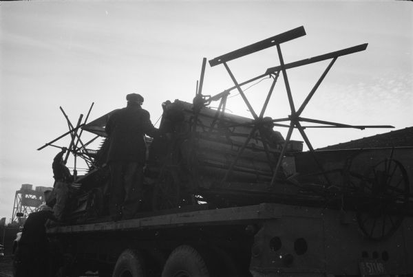 A piece of early farm equipment and two men can be seen on top of a truck bed, they are lit from behind by sunlight. Original caption: "International trucks delivering more than 200 pieces of the McCormick collection to Stonefield, Wisconsin's state farm museum."