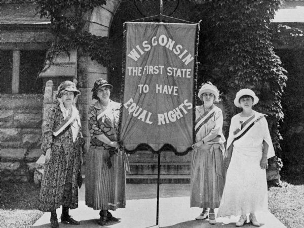 Group of Wisconsin delegates posing with banner outdoors. Left to right: Mrs. Jackowsky-Peterson, Mrs. Gustav Hipke, Mrs. Max Rotter, Miss Lenore Cawker. From the "Winning of the First Bill of Rights for American Women" by Mabel Raef Putnam.
