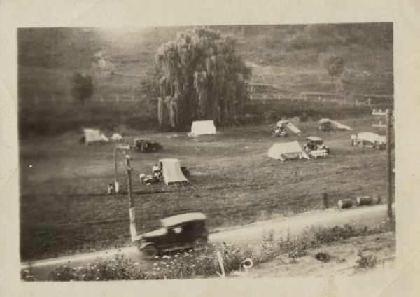 Elevated view from steep hillside towards a campsite along a road, with a large willow tree in the background behind the tents. Power poles and lines are along the road, and a car (blurred) is being driven toward the left.