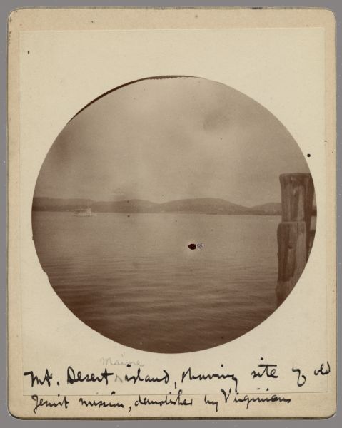Circular-framed view across a body of water. Two wood pilings are in the right foreground, and a boat is out on the water. Hills are along the far shoreline. Handwritten at bottom: "Mt. Desert Maine island, showing site of old Jesuit mission [Saint Sauveur Mission], demolished by Virginians." [Led by Captain Samuel Argall].