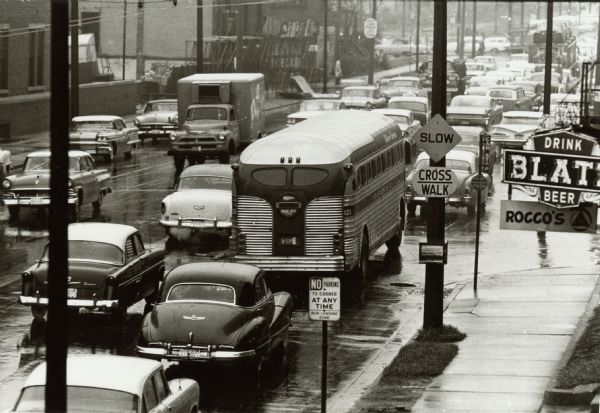 Slightly elevated view of cars, trucks, and buses in a traffic jam. A sign is on the right for Rocco's, under a neon sign to "Drink Blatz Beer."