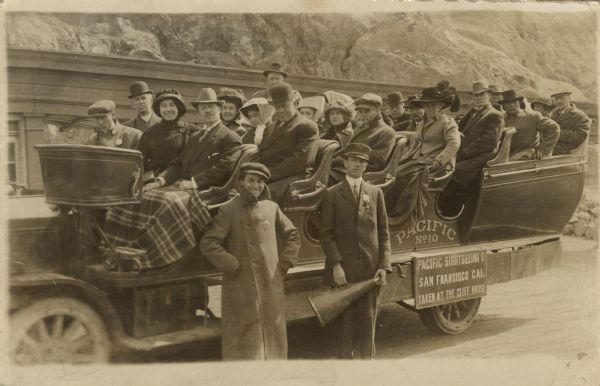 Opera group excursion in San Francisco. The group is in a Pacific Sightseeing Co. tour bus at the Cliff House. Olivia Monona is in the center of the front row of seats sharing a lap blanket with her husband Oscar Hanke. One of the tour guides is holding a megaphone.
