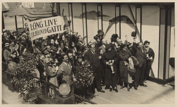 Elevated view of a crowd of Metropolitan Opera singers celebrating longtime manager of the New York Metropolitan Opera, Giulio Gatti-Casazza. The surprise celebration took place on boaerd the Italian liner <i>Rex</i> prior to Gatti's departure for Italy where he lived in retirement. Some of the performers are carrying a banner reading: "Long Live Our Beloved Gatti." Gatti is at the center of the front row with Rosa Ponselle to his left wearing a dark hat and holding a loving cup. His wife Rosina Galli is second from his right