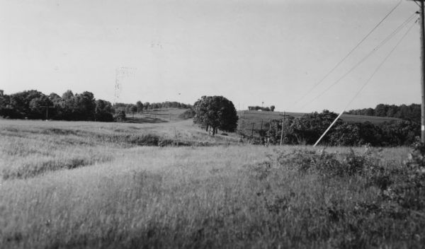 View across a gently sloping prairie field. Trees are in the distance, and power lines and a road are on the left. The Highway Commission descriptive remarks read: "Taken about (+- 1915) on Spring Green Plain Road, June 1950."