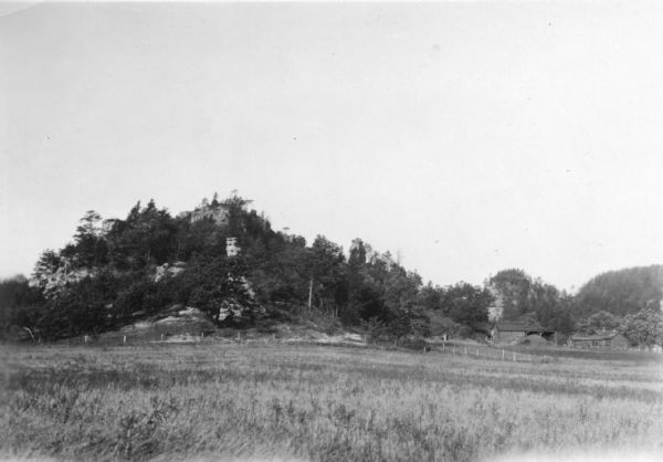 View of a rock formation with trees growing on and around it. Fence posts, power lines, and some farm buildings are nearby on the right. Caption reads: "Rock formation S.T. H 21 & 12, between Hustler and New Lisbon."
