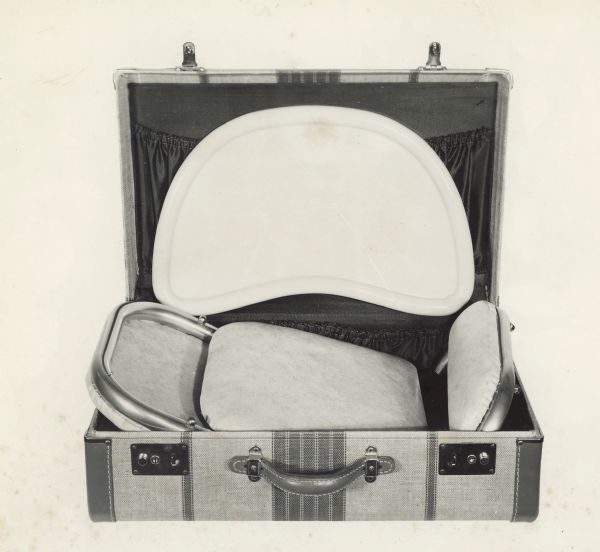 Metal baby high chair capable of disassembling and folding for easy transport. Close-up view of chair disassembled, with parts packed in a suitcase. View does not include the frame. Taken for Halbert M. Wood, WooDick Corporation.