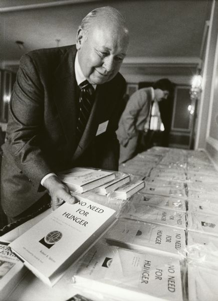 Robert Spitzer holding a book with the title: "No Need For Hunger." He is standing over a table full of the books laid out for display.