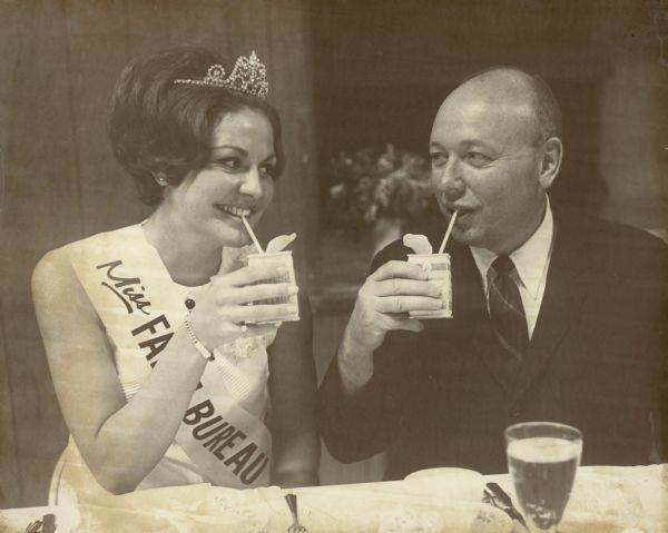 Robert Spitzer and Miss Farm Bureau sitting at a table. They are using drinking straws to drink from the containers they are holding. Miss Farm Bureau is wearing a sash and a tiara.