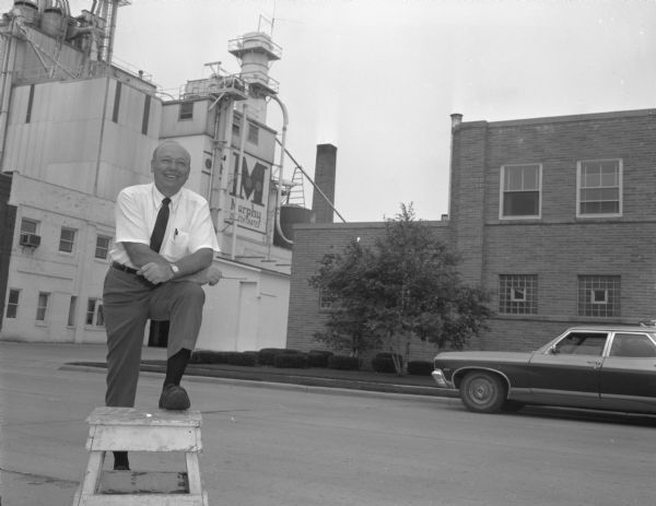 Robert Spitzer is standing outdoors with his foot up on a wooden stool. Behind him are factory buildings, with a sign that reads: "Murphy Concentrates."
