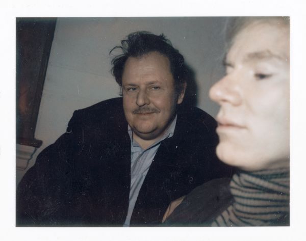 Color photograph (Polaroid) of Emile de Antonio looking at Andy Warhol during the making of the film "Painters Painting." Warhol is closest to the camera and is seen in profile. de Antonio is sitting at the center in the background.