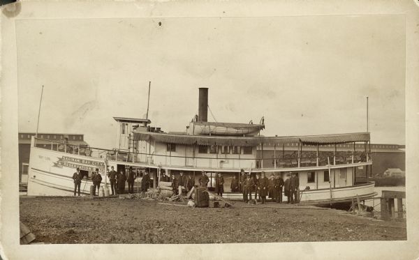 A group of people posing standing in front of the steamboat "Plowboy." Buildings are in the background.