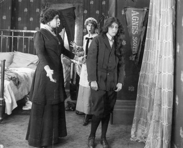 Julia Swayne Gordon, Flora Finch, and Clara Kimball Young are seen in a still from the 1913 short "When Mary Grew Up." Young is dressed as a boy but has her hair down. Gordon looks at her sternly while Finch has a look of surprise on her face.