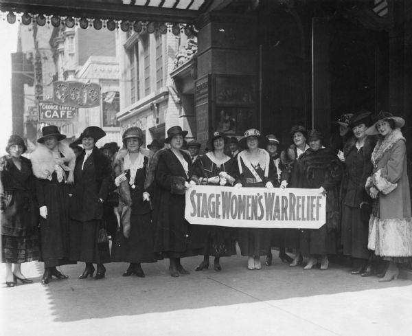 Renee Harris and other fashionably-dressed women holding a banner that reads: "Stage Women's War Relief" outside the Hudson Theatre. 