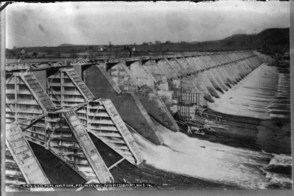An elevated view of the Gate Piers at the power dam. Men are working on top of the dam. Farm buildings and bluffs are on the opposite shoreline.