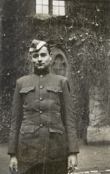 A candid portrait of John Lavalle in uniform standing in front of a vine-covered building. He is holding a cigarette in his left hand.