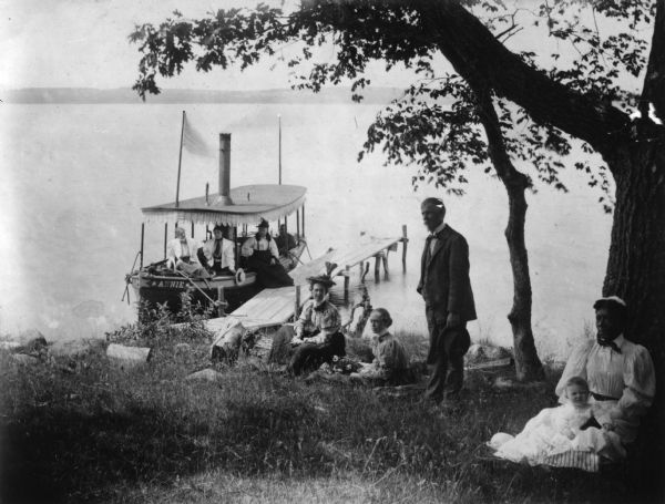 View towards shoreline with several people on or near a docked boat next to a pier. An African-American woman is sitting under a tree with a white baby on her lap. All the other people are white. A man is standing holding his hat. The boat is named "Annie." Caption reads: "Probably Green Lake, Wis. 1895-1900. From the Samuel Pedrick Albums, Ripon (Wis.) College Archives."