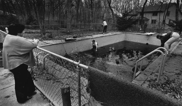 In the left foreground a woman is leaning on a fence while watching three people cleaning a half empty pool. Caption reads: "About 50 members of the Letterwinners Club at New Berlin Eisenhower High School offered a hand over the weekend by helping senior citizens with spring cleanup projects. Mary Jane Kahl watched as Tom Blair (in pool), group coordinator Tom Kelenic (right) and Tony Bolle cleaned the Kahl pool."