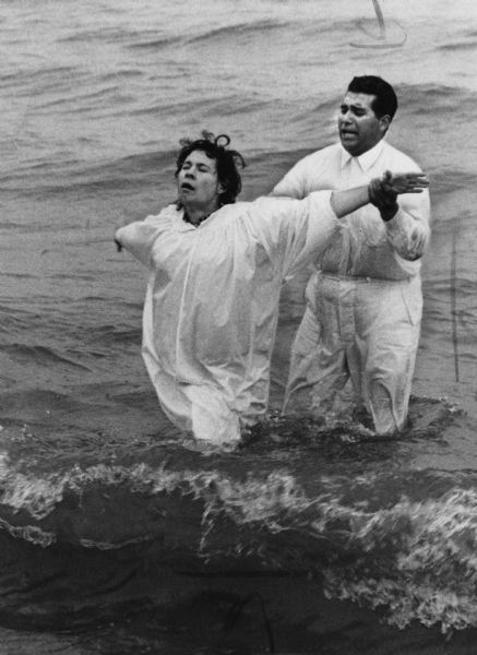 A man wearing a white shirt and pants is holding the arms of a woman who is wearing a white robe and spreading her arms out. They are both standing knee deep in water. Caption reads: "<b>The Rev. Ray E. Lopez,</b> pastor of the Apostolic Assembly of the Faith in Christ Jesus, 606 S. 5th St., baptized Mrs. Maria Martinez Saturday in Lake Michigan. Four persons were baptized in the ceremonies at Bradford beach."