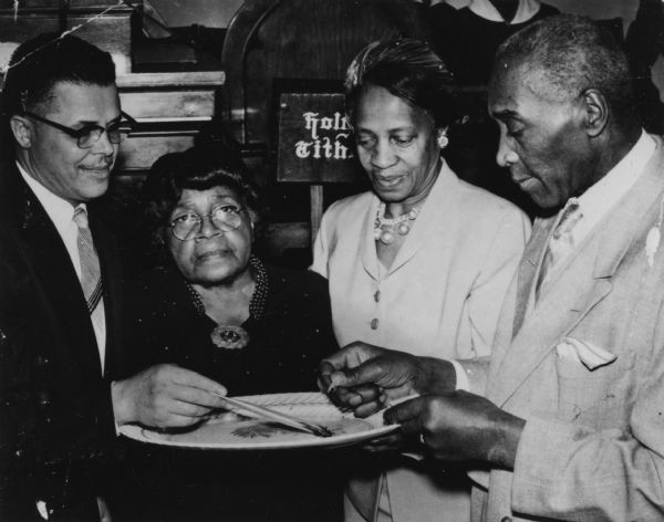 Two men and two women are standing around a plate, over which one man is holding a singed document while another man is holding a match. Caption reads: "Madison, Wis. Dec. 18, 1955. Mt. Zion Baptist Church, 548 W. Johnson St. Mortgage-burning ceremony. Left to right: Rev. Joe Dawson, Anna Hines, charter member and widow of first pastor; Mrs. Batie; George Harris."