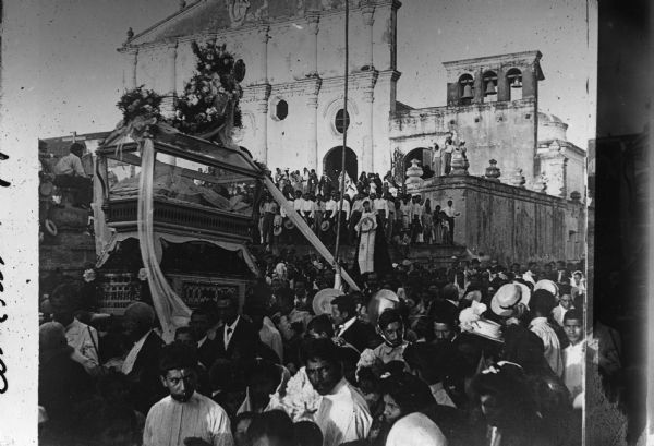 Slightly elevated view of a procession of people walking along a street past a church. They are carrying a float with a glass coffin. Flowers and streamers decorate the float.