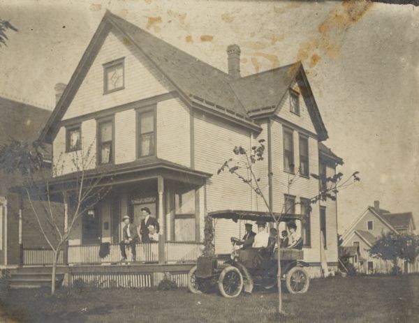 An automobile with driver and four passengers is parked beside the porch of a two-story wood frame house at 1923 East Washington Avenue. On the reverse of the photograph is written: "In car (right hand drive) (5th car in Madison) 'Pa', 'Ma', Jul, George, Dora." There are two young men and a little girl on the porch. The automobile has a solid roof with canvas sides, and there is a wicker basket over the rear fender. The automobile maker is not identified.