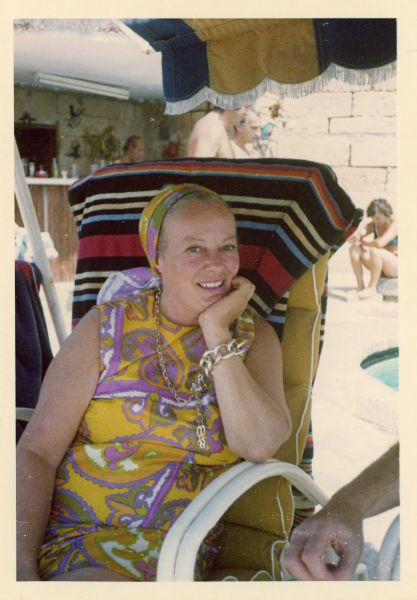 Faye Emerson siting in the shade in a chair by a pool in Majorca, Spain. She has a scarf on her head and is wearing a sleeveless yellow and purple paisley dress. She is resting her chin on her hand; her elbow is on the armrest of the chair. 