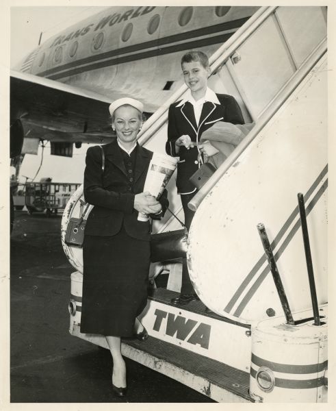 Faye Emerson and her son William "Scoop" Crawford standing on a set of stairs leading up to a TWA airplane. Faye is holding several magazines and a purse. Faye and Scoop are dressed in suits and both have cameras with them.