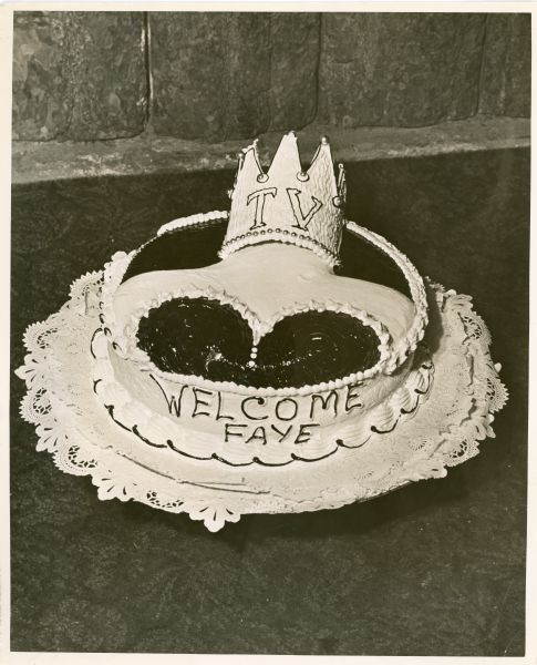 Circular cake which reads: "Welcome Faye" on the side. The top of the cake is decorated with a woman's shoulder and bust. A crown with "TV" on it is placed where someone's head would be.