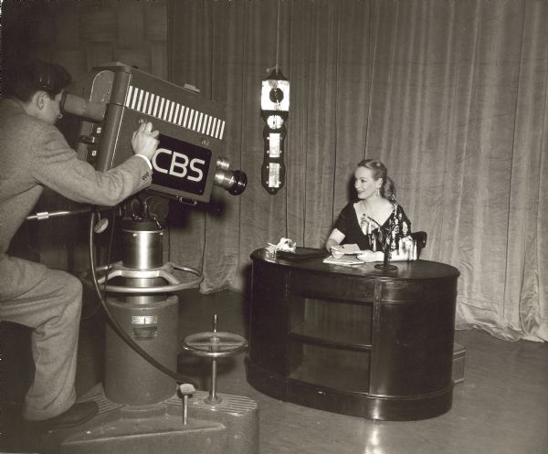 Faye Emerson sitting at a desk while filming one of the episodes of The Faye Emerson Show. Next to her on the desk is a stand with many pieces of jewelry on it. She also has several pieces of jewelry on one ear. The cameraman and CBS camera are in front of her.
