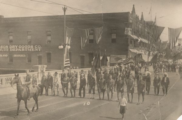 Uniformed soldiers marching in a World War I homecoming parade. Many men are carrying flags, and the street is heavily decorated with U.S. flags. The soldier in front is riding a horse. Several men not in uniform and a boy are also walking along the street. The parade is moving over a bridge. The Club billiards hall is in the background.