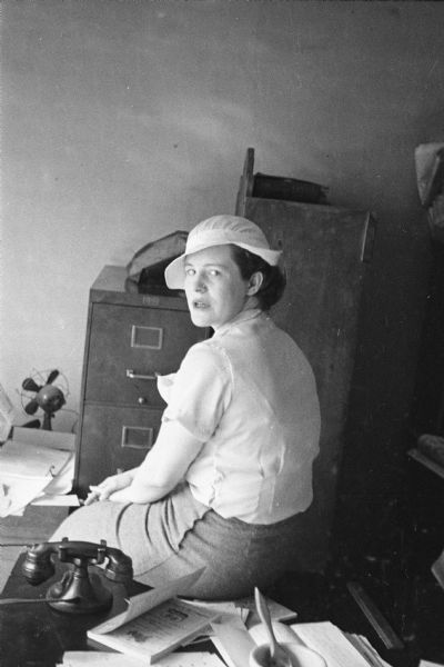 Informal portrait of Betty Cass sitting on a desk and looking over her left shoulder. On her desk is a fan, a telephone and a book to her left, and a file cabinet is behind her.