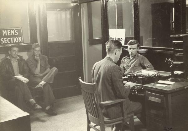 Men looking for a job during the Great Depression waiting their turn in the Employment Office waiting room, while another man is sitting with a clerk and filling out forms in hopes of finding work.