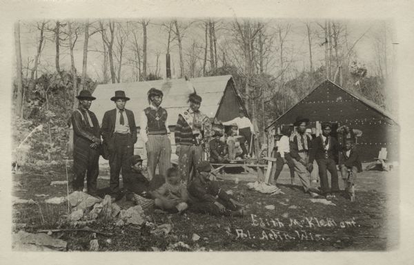 Informal outdoor group portrait of Native American men and boys. One man is wearing a feather on his head, a beaded sash, and beaded pants. Two buildings are in the background.