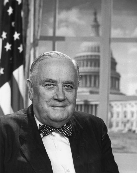 Quarter-length portrait of Senator Alexander Wiley, with a United States flag and the U.S. Capitol building in the background.