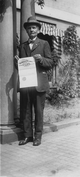 Former Wisconsin State Senator David G. James, Republican from Richland Center (1908-1912) and the father of suffragist Ada James, on his trip to Washington, D.C. to deliver Wisconsin's ratification on the 19th Amendment. He is standing outside the National Woman's Party headquarters at 14 Jackson Place. The document he is holding is possibly his appointment by Governor Phillip as Special Courier.