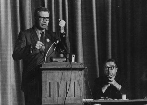 Donald O. Peterson gesturing while speaking at a podium, while Patrick J. Lucey, sitting at right, is listening. The two men were vying for the Democratic nomination for Governor.