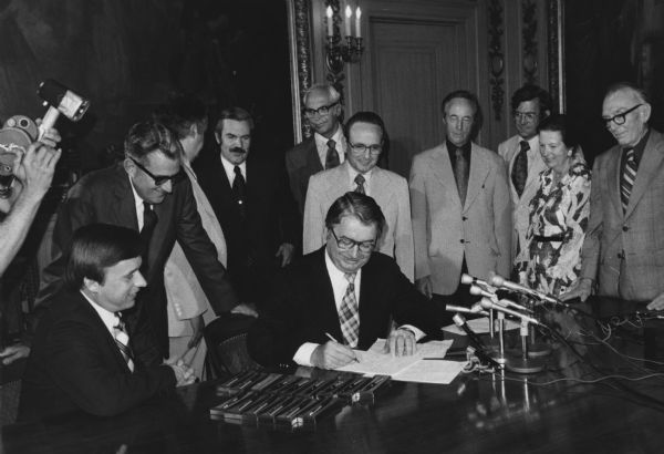 Governor Patrick J. Lucey signing the Bill merging the University of Wisconsin and the state university system, with several onlookers surrounding him. There are microphones set up on the table, and a photographer's hands holding a camera is on the left. Fifteen pens are arranged at the Governor's side for use in the signing. Future Governor Lee Dreyfus is third from the left, looking over towards another man.
