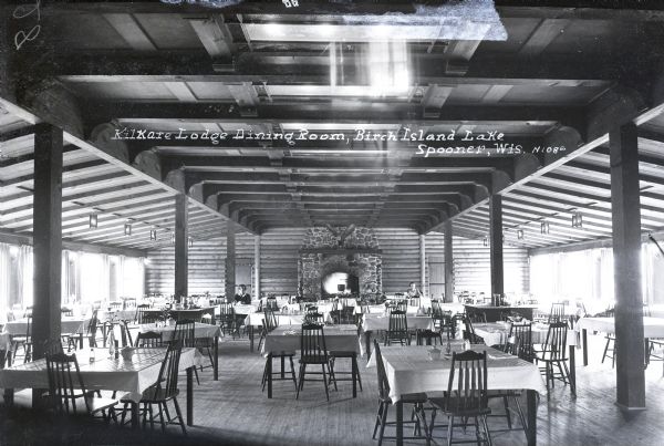 The dining room in Kilkare Lodge on Birch Island Lake. Windows line both sides of the room. Two women are sitting at tables at the far end of the room near a large, stone fireplace. A large pot is hanging from a hook at the mouth of the fireplace. On the mantel is a stuffed eagle, with wings spread.