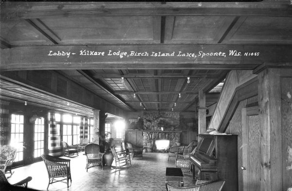 The lobby, with a stone fireplace at the Kilkare Lodge on Birch Island Lake. There are stuffed deer on mantels flanking the fireplace, and on the mantel above the fireplace is a rifle, and two stuffed birds. In the foreground on the right is a door underneath the stairs, and a piano. Windows line the wall along the left, and a stuffed deer head is mounted in between a set of doors.