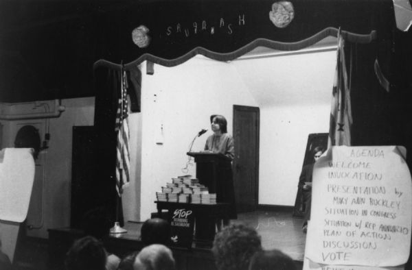 View over audience towards a woman standing on a stage in front of a podium and microphone. In front of the podium is a small table with stacks of papers or books and a sign that reads: "Stop Bombing El Salvador." A large sheet of paper on the right reads: AGENDA, WELCOME, INVOCATION, PRESENTATION by MARY ANN BUCKLEY, SITUATION IN CONGRESS, SITUATION w/ REP ANNUNZIO, PLAN OF ACTION, DISCUSSION, VOTE.
A caption on the photograph reads: "Mary Ann Buckley speaking before community meeting. 5,000 voter action cards are in front of her - they urge Rep. [Frank] Annunzio to oppose the Salvadoran air war."