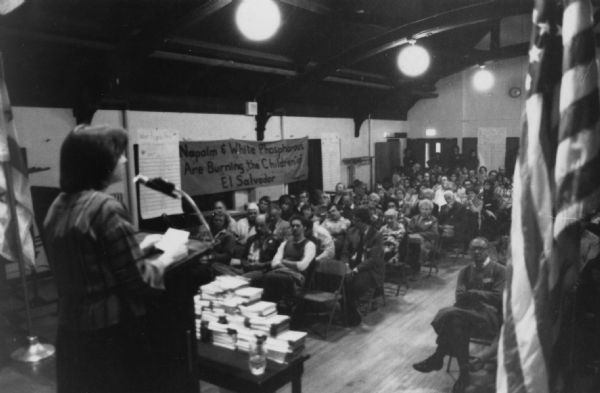 View from stage of a woman standing at a podium, with her back to the photographer. In front of her is a table stacked with cards or books, and an audience sitting on folding chairs. A banner on the far wall reads: "Napalm and White Phosphorous Are Burning the Children of El Salvador." Caption on the photograph reads: "Mary Ann Buckley, nat'l [national] CISPES coordinator, addressing crowd of 170 at community meeting about U.S.-sponsored air war, held in Rep. Frank Annunzio's NW Chicago district."