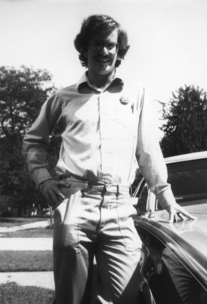 A man is standing and posing next to a car. He is wearing glasses and has a small button on his shirt that reads "Stop the Bombing in El Salvador CISPES." Caption reads: "John Adler, trainer in Chicago."