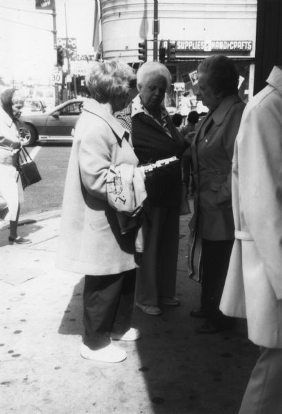 A woman is standing on a sidewalk holding something in her hands that she is showing to two other women. Caption reads: "Getting cards signed in [Representative Frank] Annunzio's district."