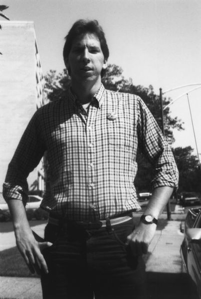 Portrait of a man standing on a sidewalk. He is wearing a wristwatch and a small button on his shirt that reads "Stop the Bombing in El Salvador CISPES." Caption reads: "Tom Goddeeris, organizer, Detroit."