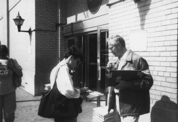 A man is standing outside a building holding a clipboard in one hand, and a pen in his other hand. A person is standing in front of him writing on a piece of paper or a folder. Caption reads: "Bob Aplanalp, volunteer in [Representative Frank] Annunzio's district, getting people to sign cards in front of Sears."